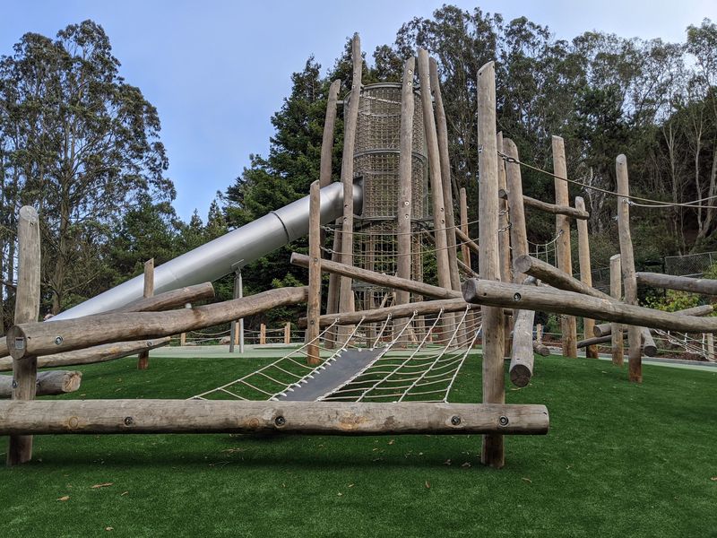 Estructura para trepar hecha de troncos y red de cuerda con una torre grande y una resbaladilla sobre una superficie de seguridad verde